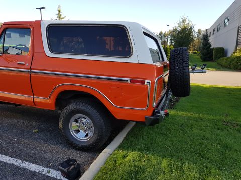 1978 Ford Bronco - The rear of an orange and white 1978 Ford Bronco is visible, parked on asphalt next to a grassy area.