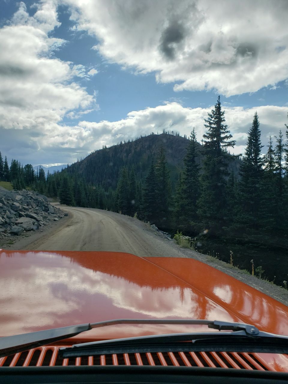 Orange vehicle hood and windshield wipers on a dirt road with trees and mountains