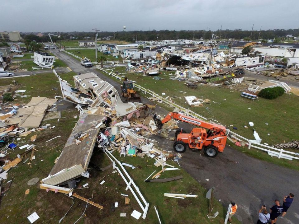 Aerial view of a neighborhood devastated by a storm, with mobile homes destroyed and construction equipment clearing debris