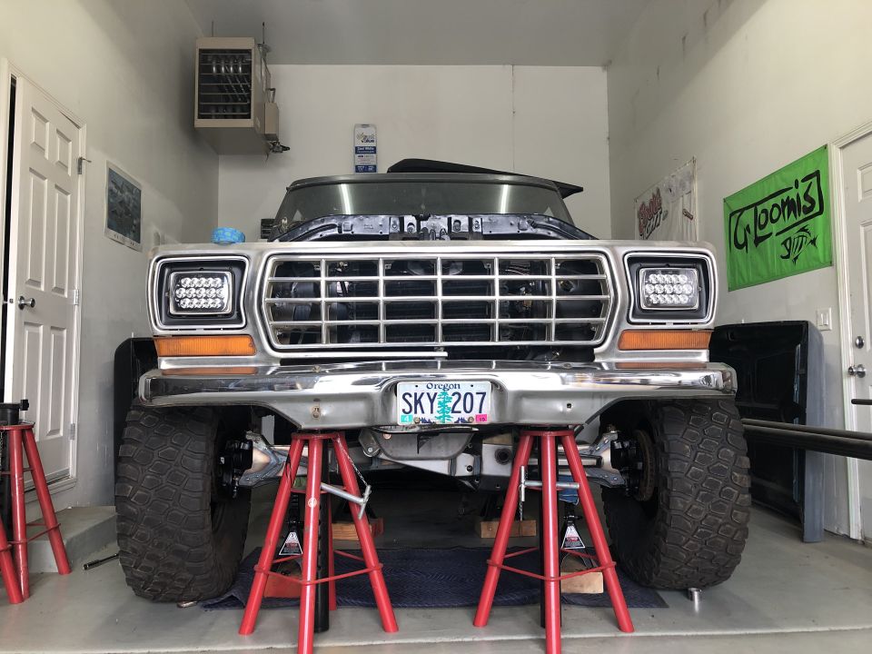 1979 Ford Bronco front end with aftermarket headlights, on jack stands in a garage. Oregon license plate SKY 207 visible