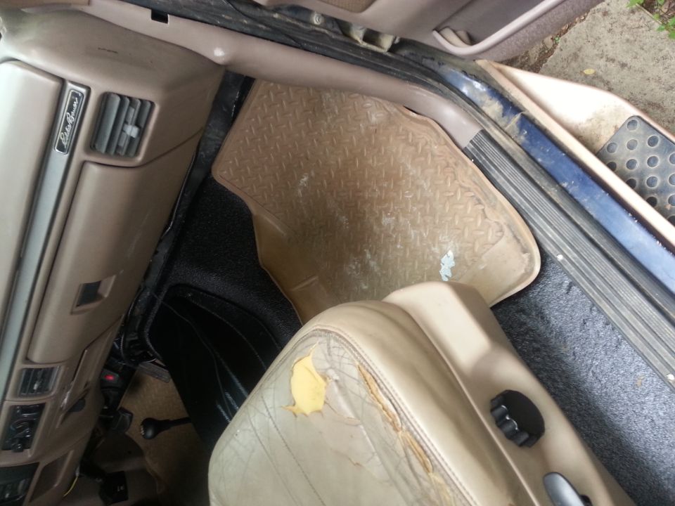 Interior of a vehicle showing a tan floor mat, torn tan leather seat, and dashboard.