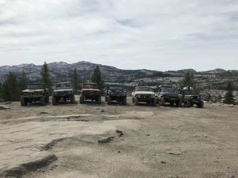 1984 Ford Bronco - Seven off-road modified vehicles are parked in a line on a rocky, dirt terrain with mountains and trees in the background.