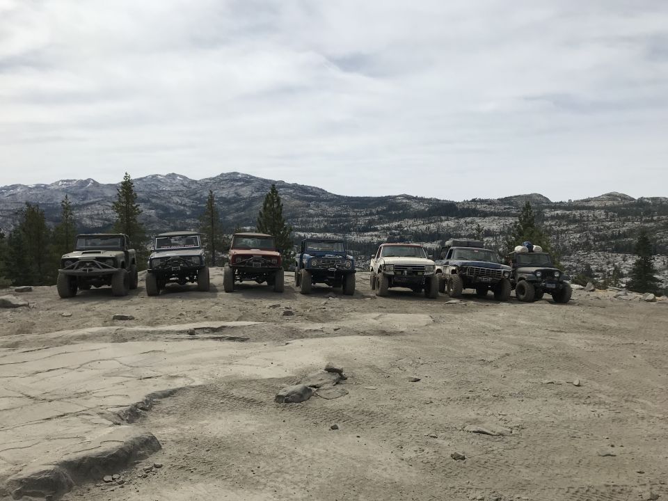 Seven off-road vehicles parked on rocky terrain with mountains in the background.