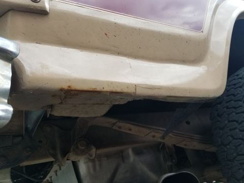 1985 Ford Bronco - A close-up view of the lower body panel and undercarriage of a vehicle, showing rust and cracking on the lower body panel. The tire and par…