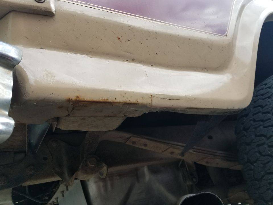 Close-up of rusted and cracked lower body panel of a vehicle, with tire and undercarriage visible.