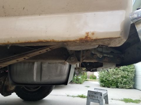 1985 Ford Bronco - The underside of a white vehicle shows significant rust along the rocker panel and frame. A fuel tank and part of a leaf spring suspension…