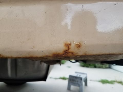 1985 Ford Bronco - A close-up view shows rust forming along the bottom edge of a cream-colored vehicle panel. Below the panel, a dark, curved object is partia…