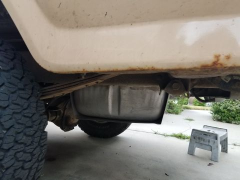 1985 Ford Bronco - The underside of a vintage vehicle shows a fuel tank mounted between the rear leaf springs and above the rear axle. Rust is visible along t…