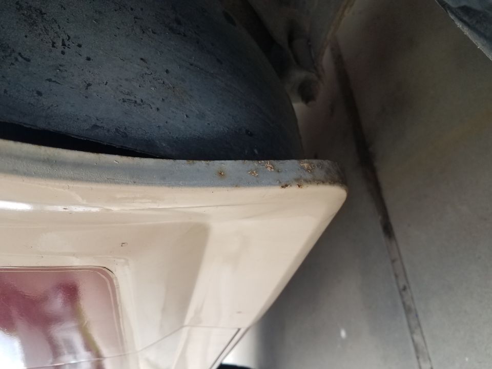 Close-up of cream-colored vehicle panel edge with rust on trim, next to a dark tire/wheel well.