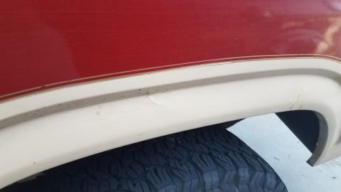 1985 Ford Bronco - A close-up view of the lower rear fender area of a red and tan vehicle, showing a tire and some minor paint chips and rust spots on the tan…