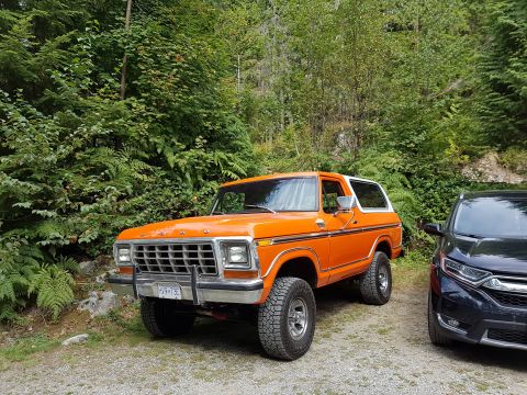 1978 Ford Bronco - A bright orange 1978 Ford Bronco is parked on a gravel road in front of lush green trees and ferns. A black SUV is parked to the right of t…