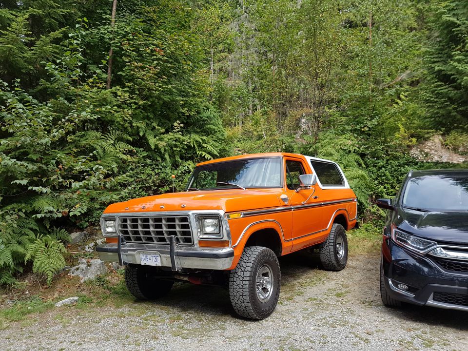 1978 Ford Bronco in orange parked on a gravel road with trees in the background.