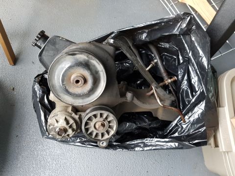 1978 Ford Bronco - A black plastic bag contains a power steering pump assembly with pulleys and hoses. The pump has a silver pulley on top and a smaller pulle…