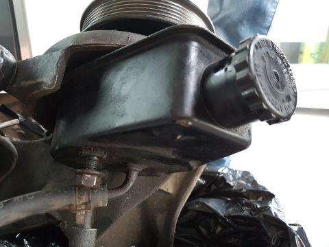 1978 Ford Bronco - A close-up, low-angle view of a black power steering pump reservoir with a ribbed black cap. A metal pulley is visible at the top, and a ho…