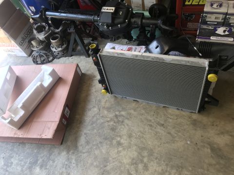 1994 Ford Bronco - A new radiator and a rear axle assembly are laid out on a concrete floor next to boxes. The radiator is silver with black plastic end tanks…