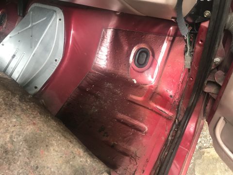 1994 Ford Bronco - The interior floor of a red vehicle, showing exposed metal with some rust and a section of insulation. A rubber grommet and wiring harness…