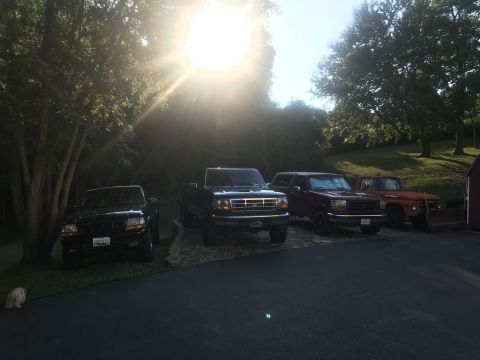 1994 Ford Bronco - Four pickup trucks and a Bronco are parked in a driveway with trees and sunlight in the background. The trucks are a black Ford Ranger, a b…