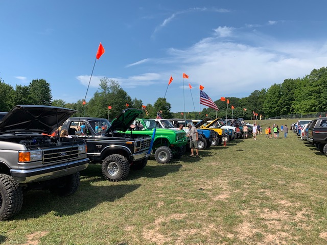 Line of lifted Ford Broncos and off-road vehicles parked on grass with hoods open and orange flags.