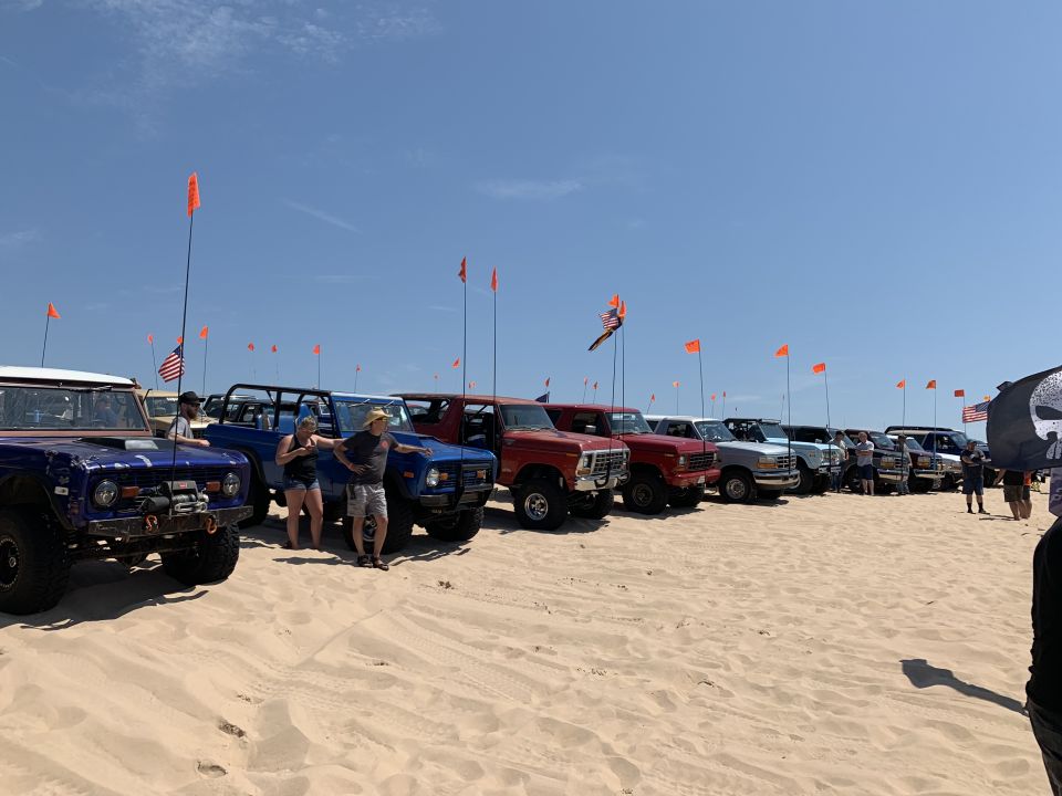 A line of Ford Broncos and trucks parked on a sandy beach with people and flags.