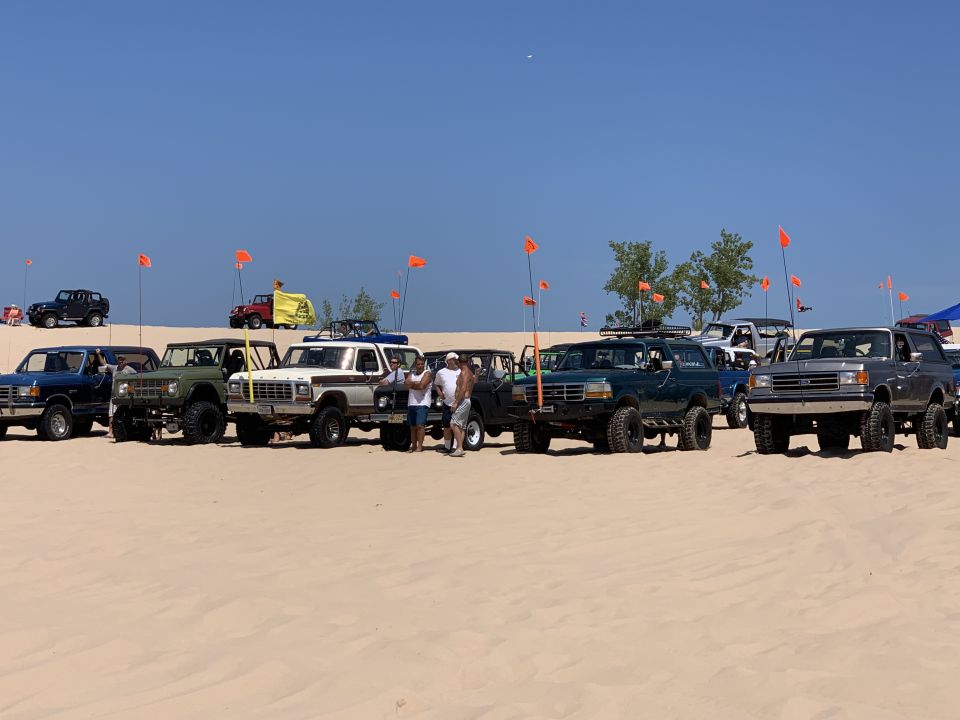 Several Ford Broncos and a Jeep parked on a sandy dune with orange safety flags.