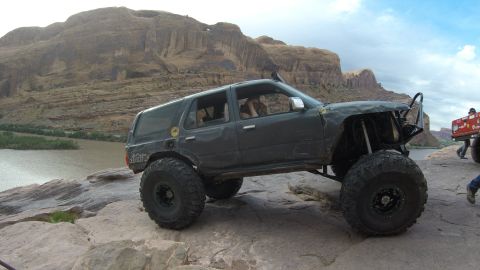 1984 Ford Bronco - A modified dark gray SUV with oversized tires is navigating a rocky, off-road terrain next to a river with a large mesa in the background.