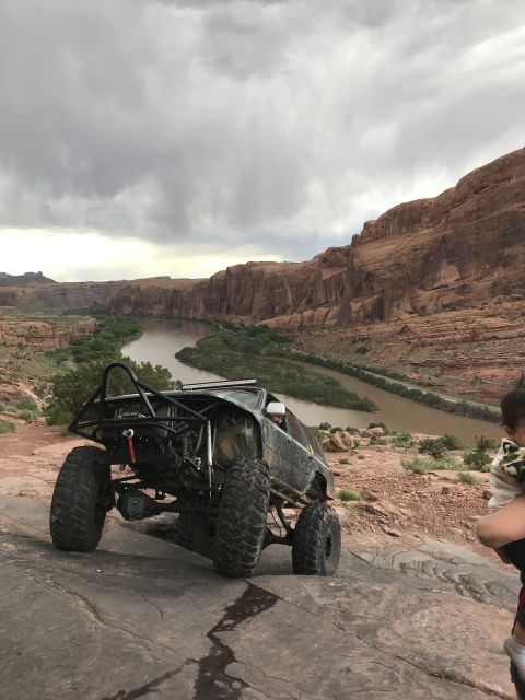 1984 Ford Bronco - A heavily modified off-road vehicle with large tires and a roll cage is positioned on a rocky incline overlooking a river valley with red r…