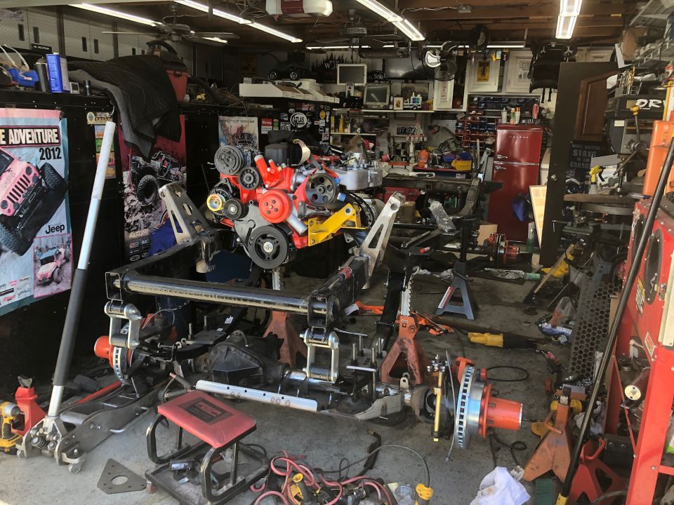 Jeep Wrangler chassis with engine and drivetrain components disassembled in a cluttered garage. Poster for "Adventure 2012" visible