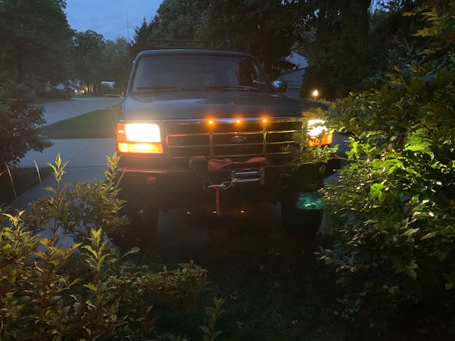 Front view of a dark Ford Bronco with headlights on, parked on a driveway at dusk.