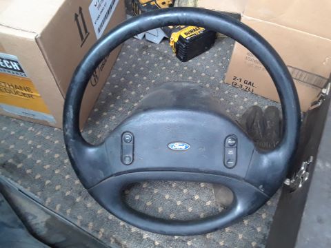 1994 Ford Bronco - A black Ford steering wheel is shown in a close-up shot. The steering wheel has a Ford logo in the center and buttons on the sides, with a…