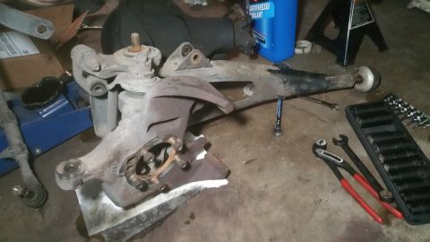 1996 Ford Bronco - Various automotive suspension parts are shown disassembled on a concrete surface, alongside a set of tools and a bottle of coolant.