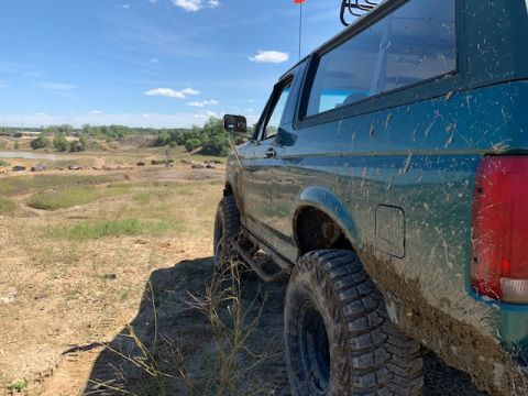 1996 Ford Bronco - A muddy 1996 Ford Bronco sits off-road under a bright blue sky, with other vehicles visible in the distance.