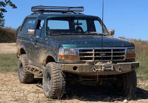 1996 Ford Bronco - Mud-covered 1996 Ford Bronco with a roof rack and winch is parked on a dirt road under a clear blue sky.