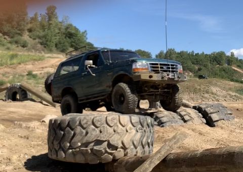 1996 Ford Bronco - A green 1996 Ford Bronco is shown off-roading, climbing over large tires on a sunny day.