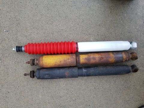 1978 Ford Bronco - Three shock absorbers are laid out on a concrete surface. The top shock absorber has a red accordion boot and a white body, while the other…