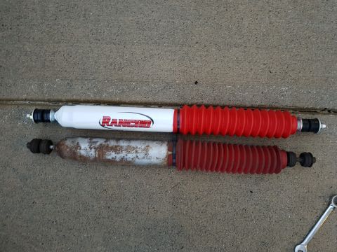 1978 Ford Bronco - A new white Rancho brand shock absorber with a red boot is shown next to an older, rusty shock absorber with a faded red boot. Both shocks…