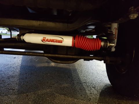 1978 Ford Bronco - The underside of a vehicle shows a white Rancho steering stabilizer with a red accordion boot installed on the steering linkage.