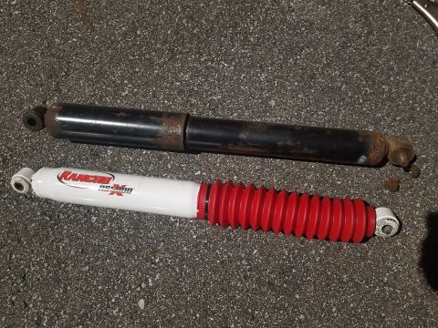 1978 Ford Bronco - A new white and red Rancho shock absorber is shown below a rusty black shock absorber, both lying on a dark asphalt surface.