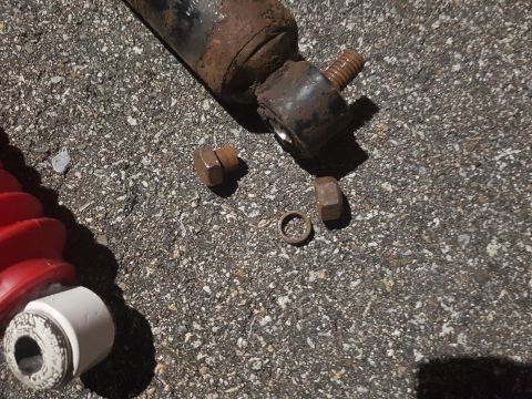 1978 Ford Bronco - A rusty shock absorber assembly with a threaded stud and two nuts, along with a washer and a separate nut, are laid out on a dark, pebbly s…