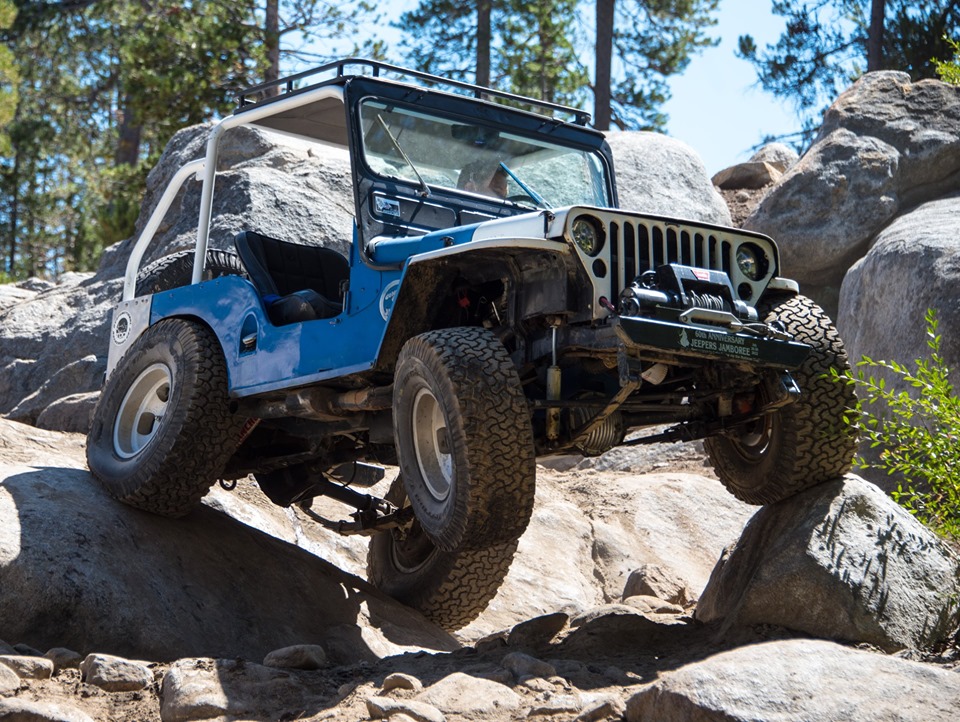 Blue and white Jeep Wrangler off-roading over rocks, showing winch and roll cage.