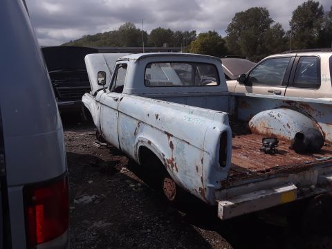 1994 Ford Bronco - A light blue, rusty pickup truck is parked in a junkyard with its hood open. Another vehicle, possibly an SUV, is parked behind it.