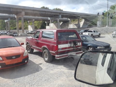 1994 Ford Bronco - A red 1994 Ford Bronco is parked on a gravel lot under a concrete overpass. A Kia Rio is parked in front of the Bronco.