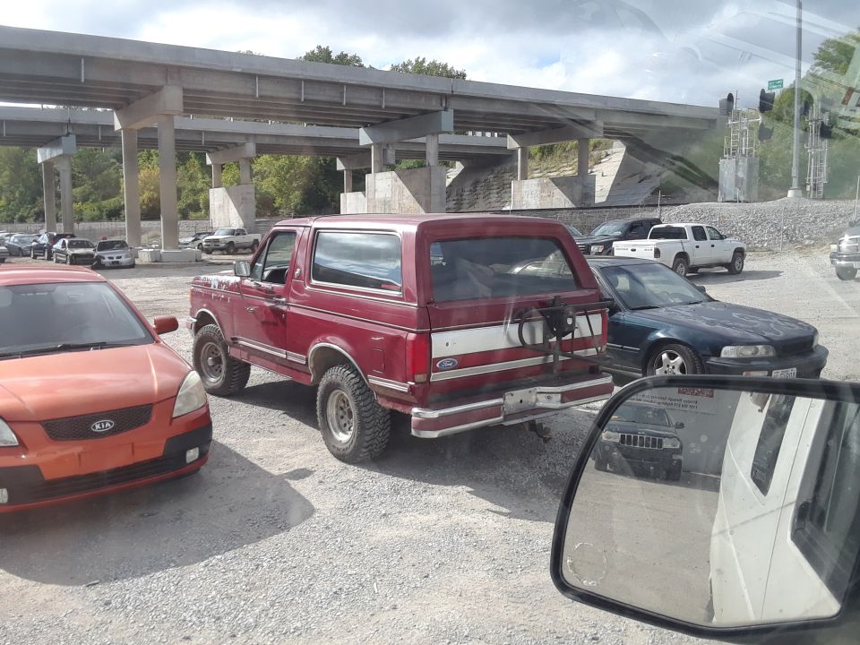 Red 1994 Ford Bronco parked on gravel under overpass with Kia Rio in front.