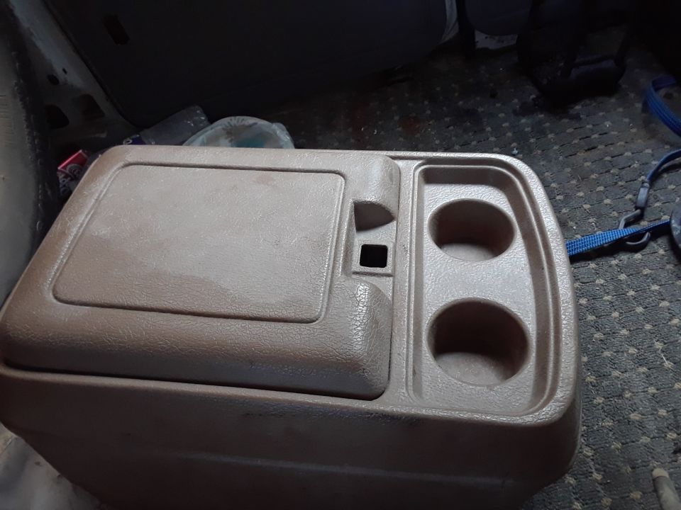 Tan plastic center console with cup holders and lid in a vehicle interior.