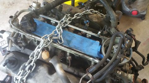 1996 Ford Bronco - A close-up view of a V8 engine with a metal chain draped over the intake manifold, which is covered in blue tape. Various hoses and electri…