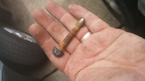 1996 Ford Bronco - A hand holds a heavily rusted bolt with a hexagonal head and threaded shaft. A portion of a tire and wheel is visible in the background.