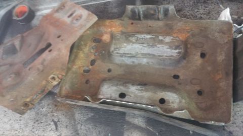 1996 Ford Bronco - Two rusty metal parts, likely from a vehicle's undercarriage, are shown on a textured surface. The larger part is a stamped metal component…
