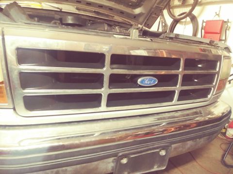 1996 Ford Bronco - A close-up, low-angle front view of a chrome grille on a Ford Bronco, featuring the blue Ford oval emblem. The hood is open, revealing part…