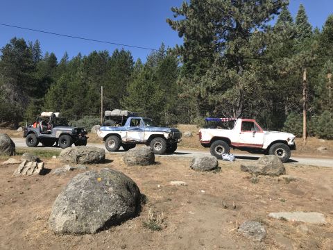 1984 Ford Bronco - Three off-road vehicles are parked on a dirt road with a forest in the background. A grey Jeep is on the left, a blue and white pickup truc…