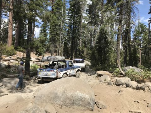 1984 Ford Bronco - A blue and white Ford Bronco is parked on a rocky, dirt trail in a forest. A person in a blue shirt and jeans stands near the front of the…