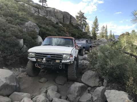 1984 Ford Bronco - A white Ford Bronco with a red roof is navigating a rocky, off-road trail, followed by a blue Ford Bronco. The white Bronco has a winch mou…